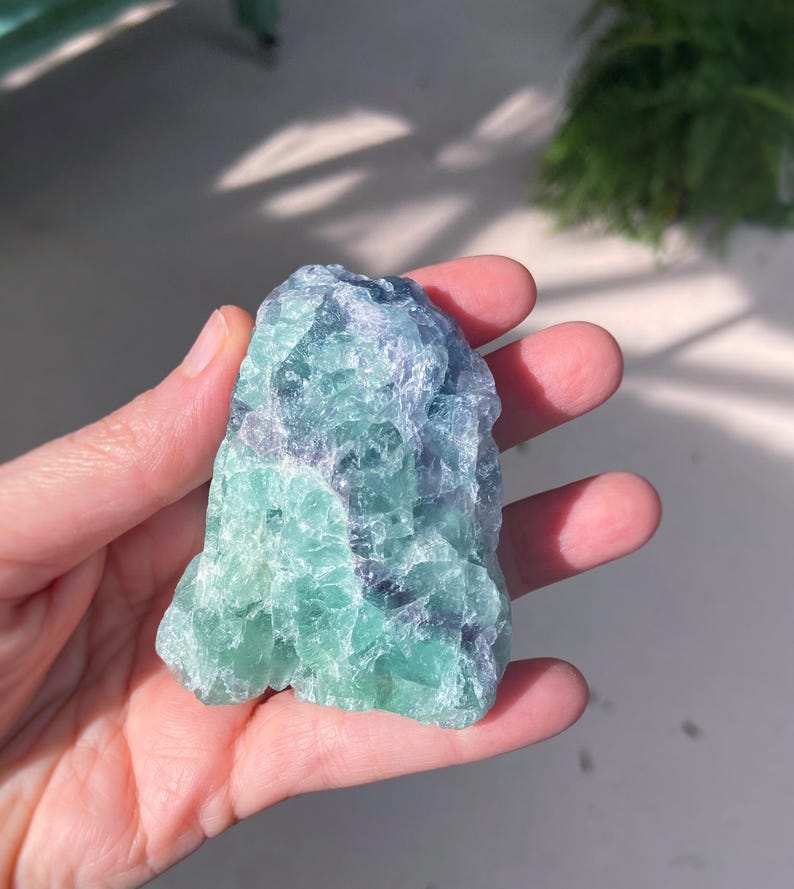 May include: A raw, natural fluorite crystal in shades of green and blue. The stone is held in a hand, showcasing its rough, textured surface and natural variations. The crystal's colors blend together, creating a visually appealing mineral specimen.