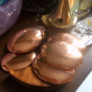 May include: Five smooth, oval-shaped copper plates, stacked on top of each other, on a wooden surface.