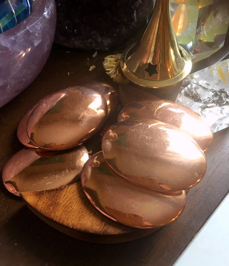 May include: Five smooth, oval-shaped copper plates, stacked on top of each other, on a wooden surface.