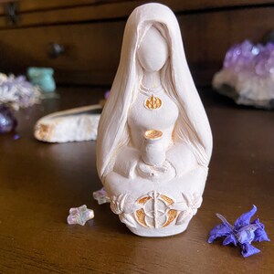 May include: White ceramic figurine of a woman with long hair, wearing a robe and holding a chalice. The figurine has gold accents and is sitting on a base with a circular design.