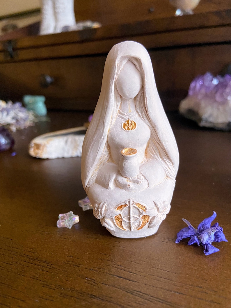 May include: White ceramic figurine of a woman with long hair, wearing a robe and holding a chalice. The figurine has gold accents and is sitting on a base with a circular design.