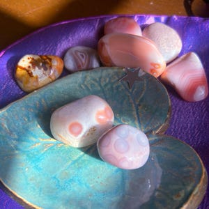 May include: A collection of polished gemstones in shades of pink, white, and brown, resting in a teal ceramic dish with a gold rim. The gemstones vary in size and shape, with some displaying banded patterns.