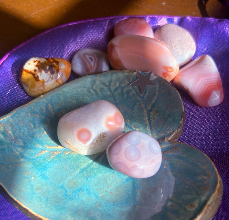 May include: A collection of polished gemstones in shades of pink, white, and brown, resting in a teal ceramic dish with a gold rim. The gemstones vary in size and shape, with some displaying banded patterns.