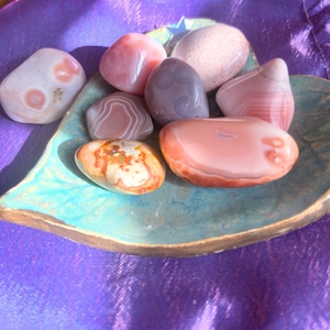 May include: A collection of polished stones in shades of pink, gray, and orange, resting in a turquoise ceramic dish. The stones vary in size and pattern, with some displaying banded designs. The dish sits on a purple, textured fabric.
