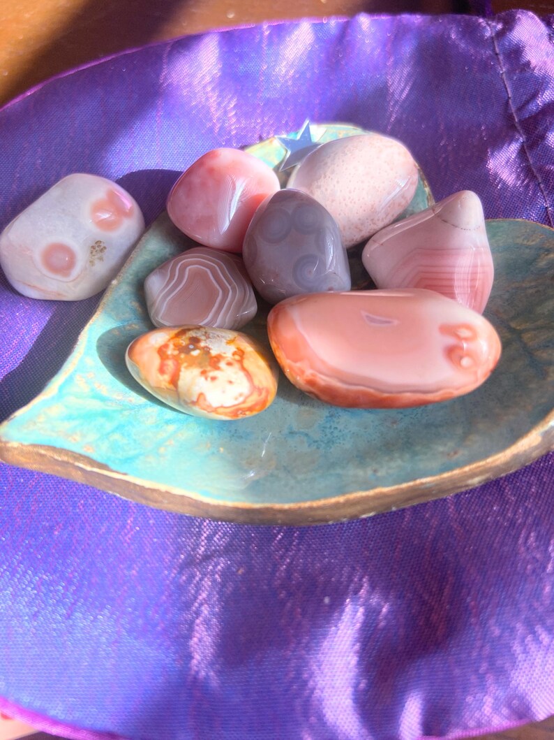 May include: A collection of polished stones in shades of pink, gray, and orange, resting in a turquoise ceramic dish. The stones vary in size and pattern, with some displaying banded designs. The dish sits on a purple, textured fabric.