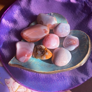 May include: A collection of polished gemstones in shades of pink, peach, and cream, resting on a teal-colored, leaf-shaped dish. The stones vary in size and pattern, with some displaying banded or speckled designs. The dish sits on a purple satin surface.