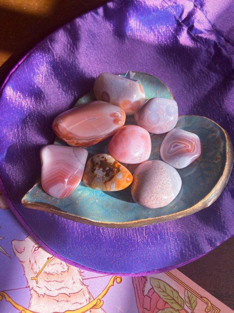 May include: A collection of polished gemstones in shades of pink, peach, and cream, resting on a teal-colored, leaf-shaped dish. The stones vary in size and pattern, with some displaying banded or speckled designs. The dish sits on a purple satin surface.