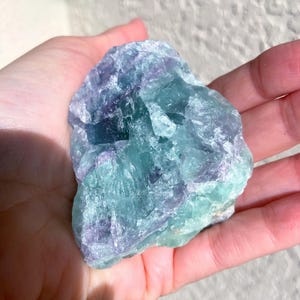 May include: A raw, natural fluorite crystal with a mix of blue, green, and purple hues. The stone is held in a hand, showcasing its rough, textured surface and crystalline structure. The colors blend together, creating a visually appealing mineral specimen.