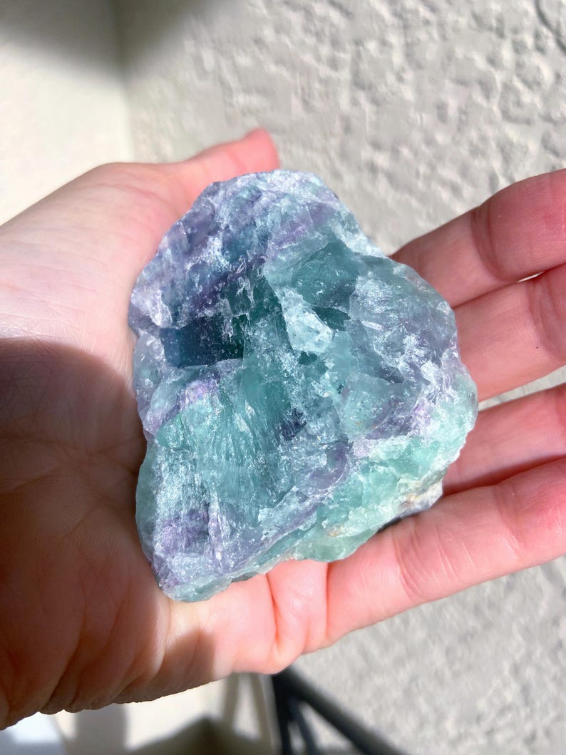 May include: A raw, natural fluorite crystal with a mix of blue, green, and purple hues. The stone is held in a hand, showcasing its rough, textured surface and crystalline structure. The colors blend together, creating a visually appealing mineral specimen.
