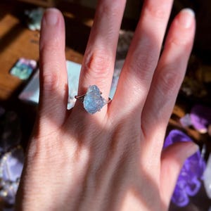 May include: A ring featuring a raw, light blue, translucent gemstone set in a polished silver-toned metal band. The stone has an irregular, jagged shape with a crystalline texture, and the band splits into two thin, delicate arms at the setting.