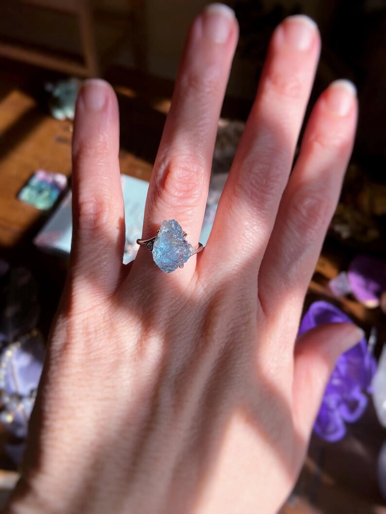 May include: A ring featuring a raw, light blue, translucent gemstone set in a polished silver-toned metal band. The stone has an irregular, jagged shape with a crystalline texture, and the band splits into two thin, delicate arms at the setting.