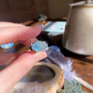 May include: A ring featuring a raw, light blue, translucent gemstone set on a thin, silver-colored metal band. The stone has an irregular, jagged shape with a crystalline texture, reflecting light across its uneven surface while held in place by a small metal prong setting.