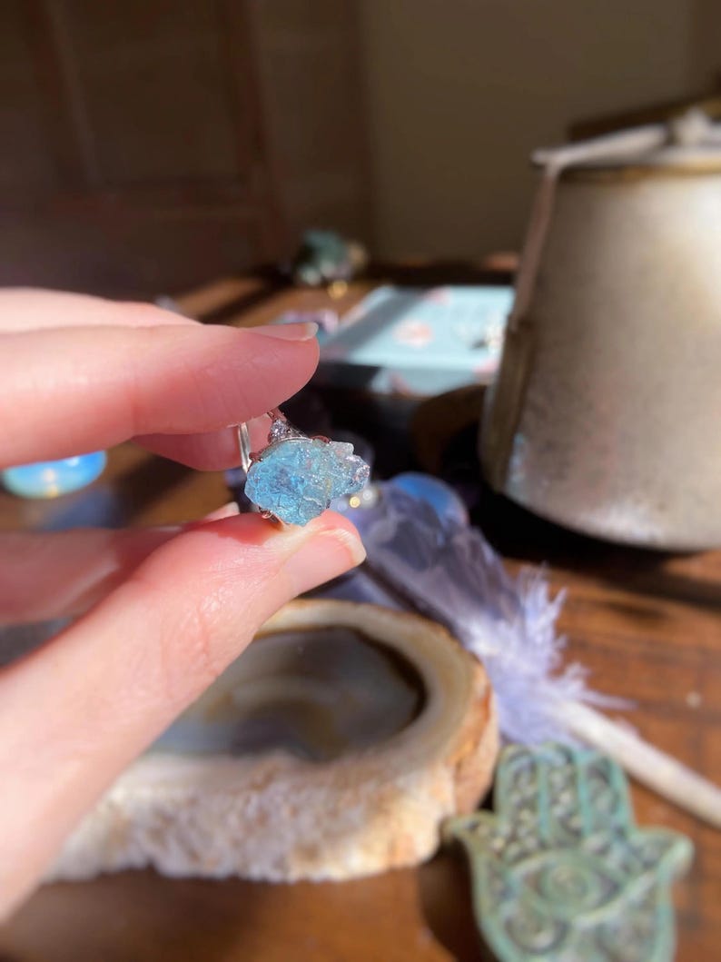 May include: A ring featuring a raw, light blue, translucent gemstone set on a thin, silver-colored metal band. The stone has an irregular, jagged shape with a crystalline texture, reflecting light across its uneven surface while held in place by a small metal prong setting.