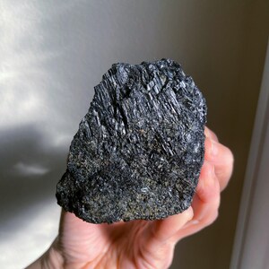 May include: A rough, black tourmaline crystal with a textured surface. The crystal is held in a hand, showcasing its dark, reflective facets and natural, raw form. The mineral appears to be a solid, dark mass.