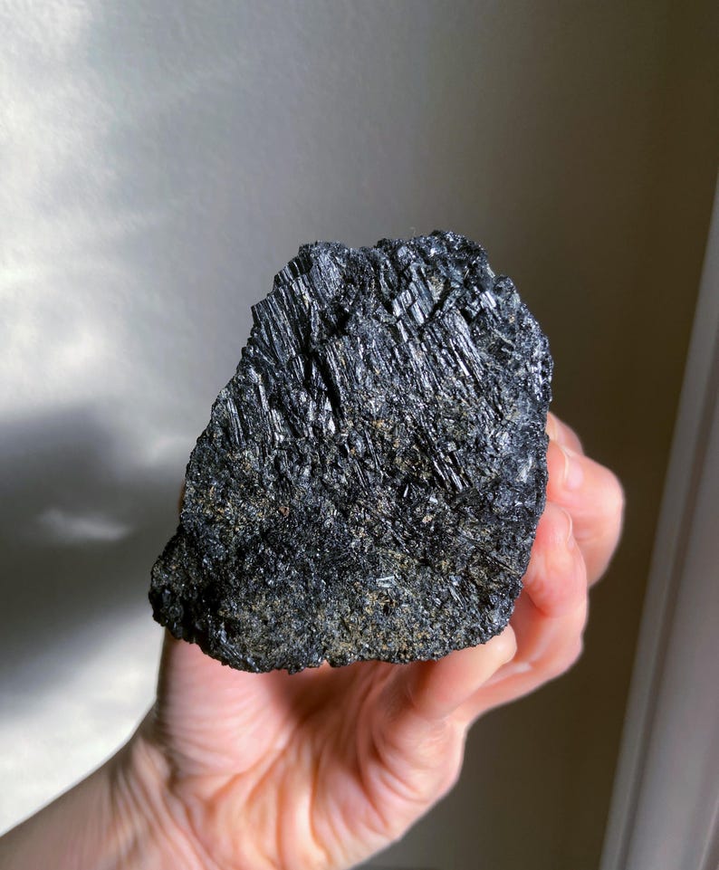 May include: A rough, black tourmaline crystal with a textured surface. The crystal is held in a hand, showcasing its dark, reflective facets and natural, raw form. The mineral appears to be a solid, dark mass.