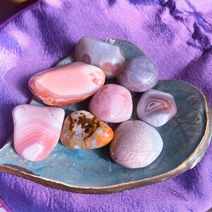 May include: A collection of polished gemstones in shades of pink, white, and brown, arranged on a teal, leaf-shaped dish. The stones vary in size and pattern, with some displaying banded layers. The background is a purple fabric.