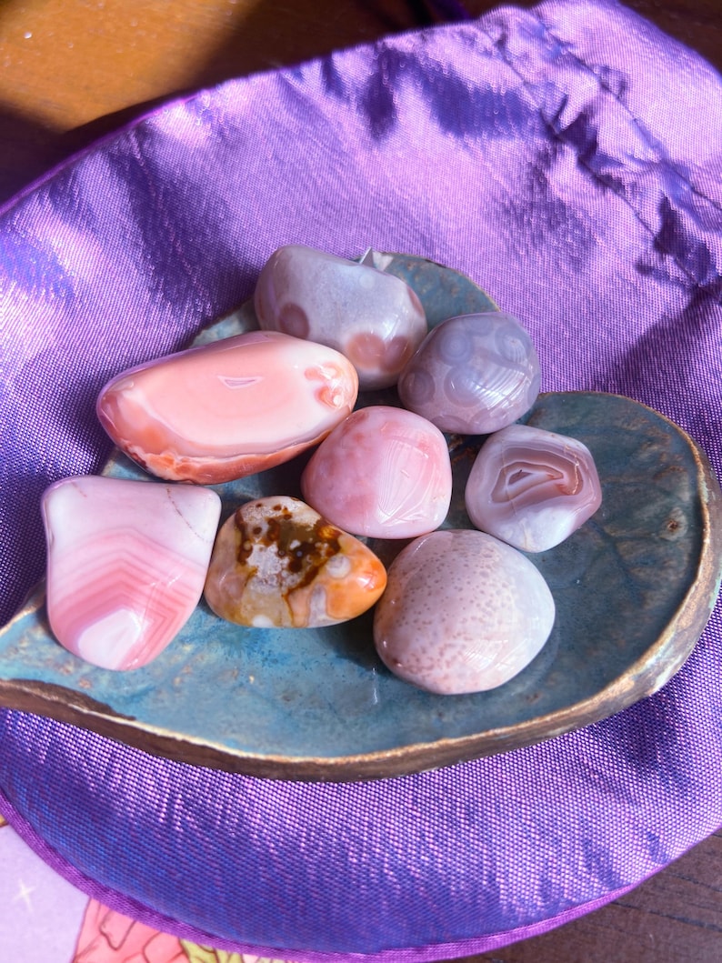 May include: A collection of polished gemstones in shades of pink, white, and brown, arranged on a teal, leaf-shaped dish. The stones vary in size and pattern, with some displaying banded layers. The background is a purple fabric.