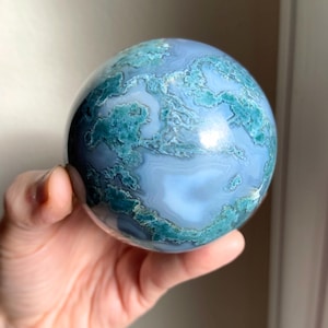 Pu&ograve; includere: Una gemma sferica lucidata, con una base azzurra e intricati motivi turchese e bianchi. La sfera &egrave; tenuta in una mano, mostrando la sua superficie liscia e le sue variazioni naturali.