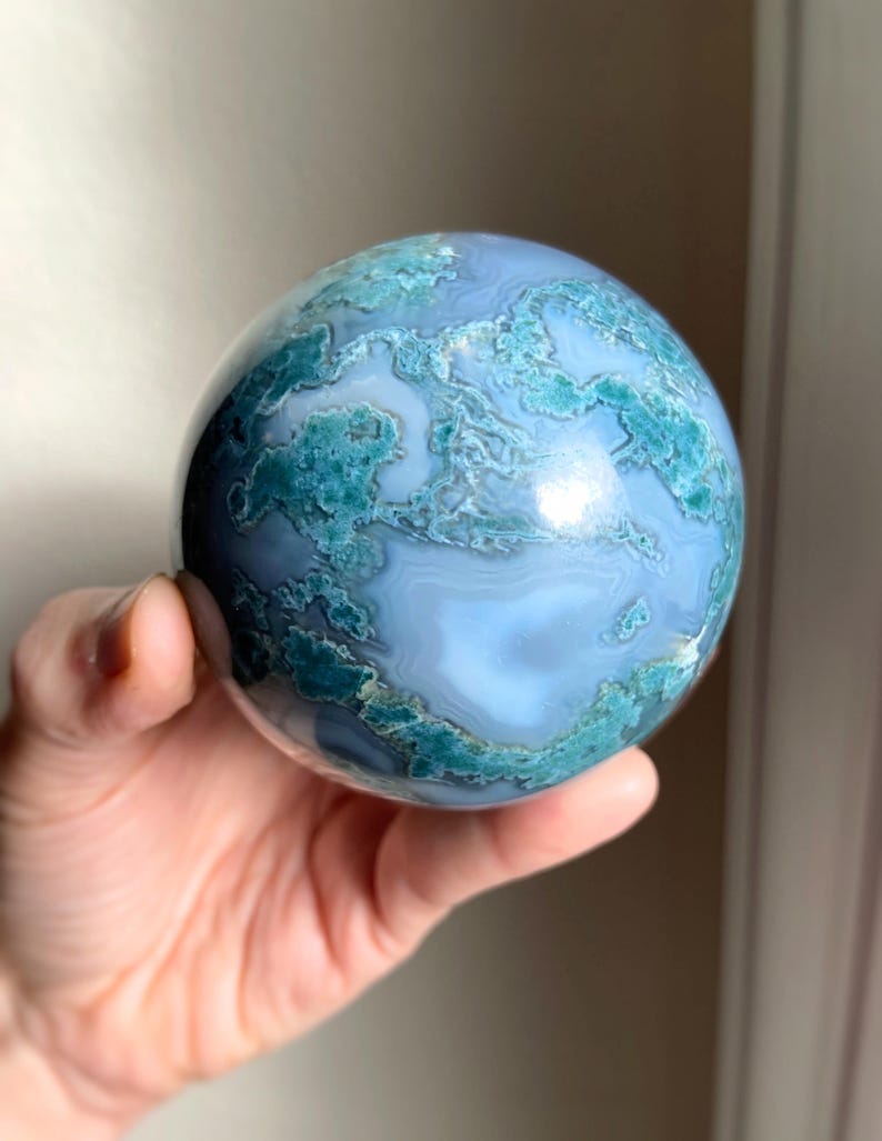 Pu&ograve; includere: Una gemma sferica lucidata, con una base azzurra e intricati motivi turchese e bianchi. La sfera &egrave; tenuta in una mano, mostrando la sua superficie liscia e le sue variazioni naturali.