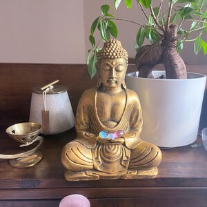 May include: A gold-colored Buddha statue sitting in meditation with three iridescent glass orbs in its hands. The statue is on a wooden surface with a potted plant and other decorative items.
