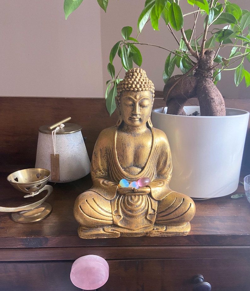 May include: A gold-colored Buddha statue sitting in meditation with three iridescent glass orbs in its hands. The statue is on a wooden surface with a potted plant and other decorative items.