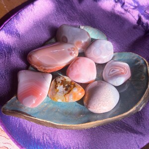 May include: A collection of polished gemstones in shades of pink, orange, and white, resting on a teal, leaf-shaped dish. The stones vary in size and pattern, displayed on a purple satin surface.