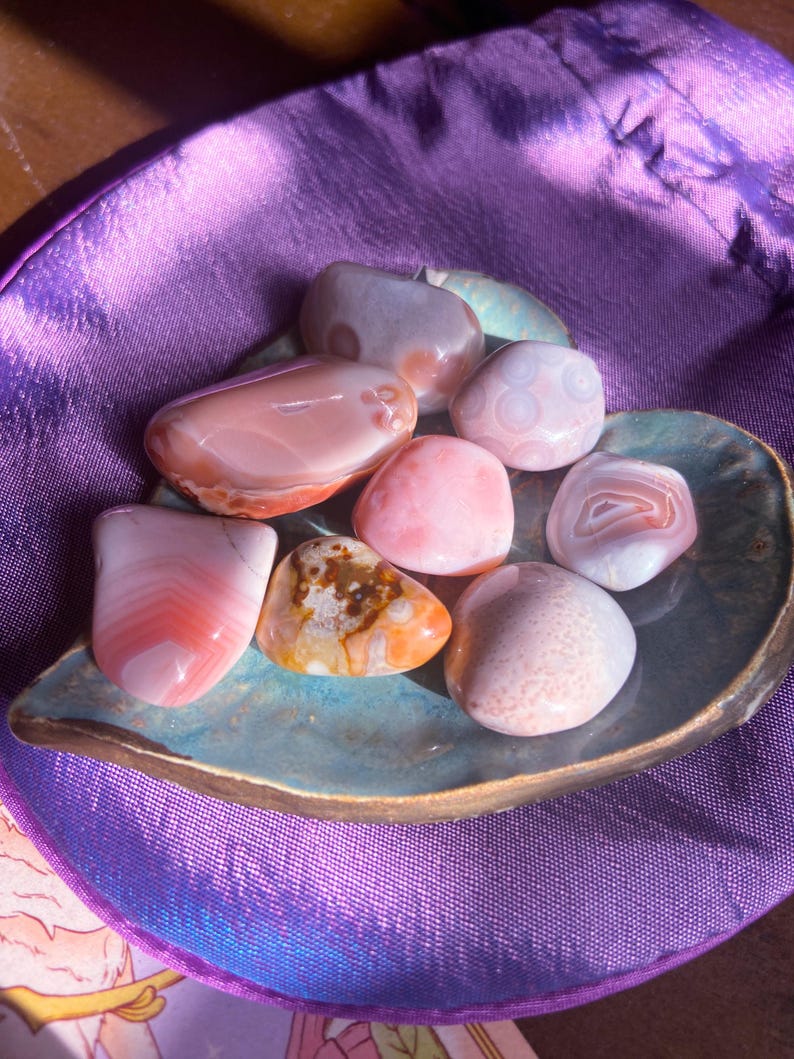 May include: A collection of polished gemstones in shades of pink, orange, and white, resting on a teal, leaf-shaped dish. The stones vary in size and pattern, displayed on a purple satin surface.