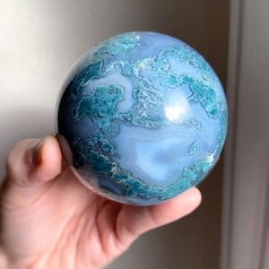 Pu&ograve; includere: Una sfera di pietra preziosa lucidata, con un motivo a vortice di tonalit&agrave; blu e turchese. La sfera &egrave; tenuta in una mano, rivelando la sua superficie liscia e riflettente. Il design della pietra ricorda un pianeta in miniatura.