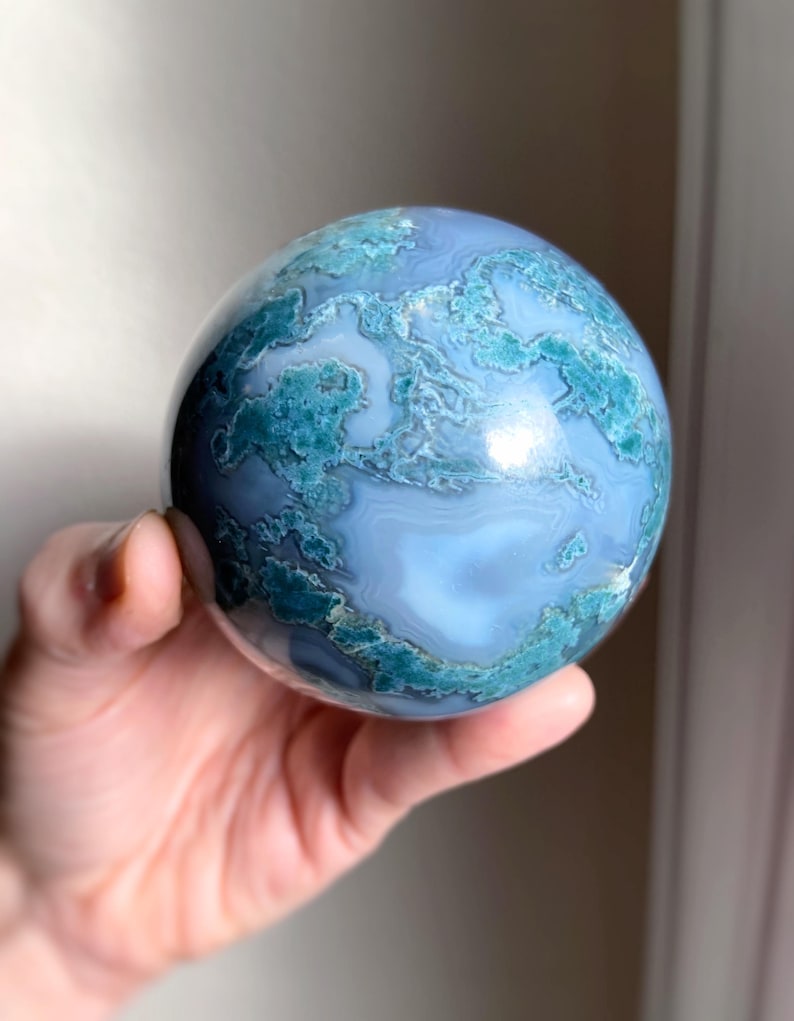 Pu&ograve; includere: Una sfera di pietra preziosa lucidata, con un motivo a vortice di tonalit&agrave; blu e turchese. La sfera &egrave; tenuta in una mano, rivelando la sua superficie liscia e riflettente. Il design della pietra ricorda un pianeta in miniatura.