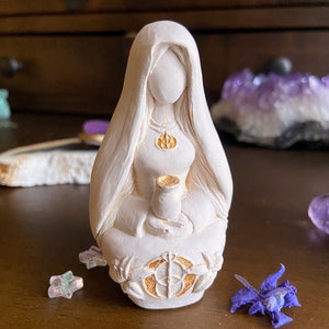 May include: White ceramic figurine of a woman sitting with her legs crossed, holding a small cup. She is wearing a long robe with gold accents and a gold necklace with a symbol. The figurine is sitting on a circular base with a gold design.
