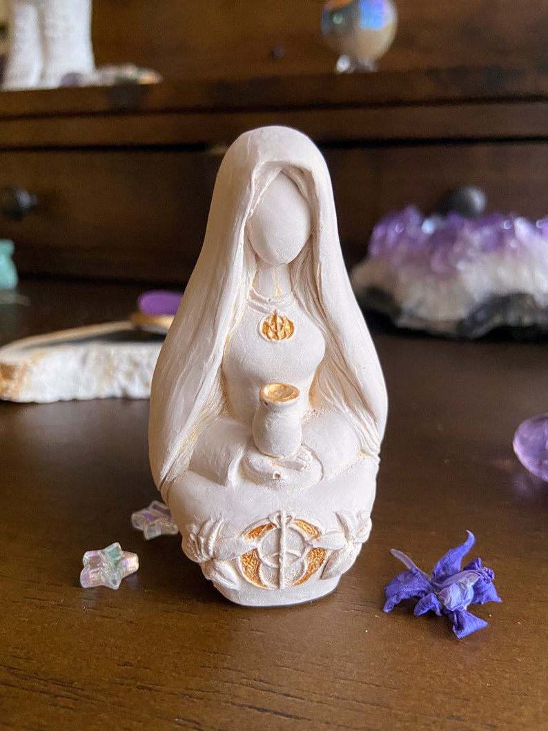 May include: White ceramic figurine of a woman sitting with her legs crossed, holding a small cup. She is wearing a long robe with gold accents and a gold necklace with a symbol. The figurine is sitting on a circular base with a gold design.