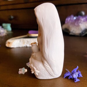 May include: White ceramic figurine of a woman wearing a long robe, holding a small pitcher in her hand. The figurine is sitting on a wooden surface.