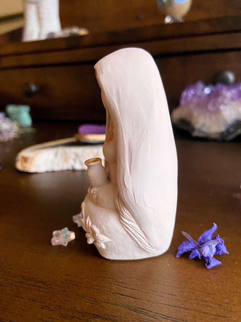 May include: White ceramic figurine of a woman wearing a long robe, holding a small pitcher in her hand. The figurine is sitting on a wooden surface.