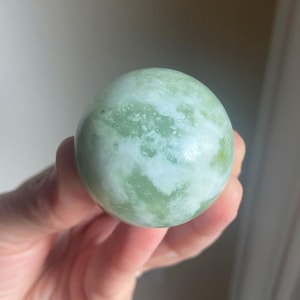 May include: A smooth, spherical jade stone. The polished surface displays a mix of pale green and white hues, with a marbled appearance. The stone is held in a hand, showcasing its size and texture. This decorative item is a natural stone.