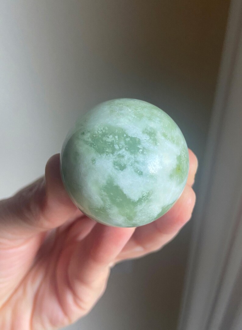 May include: A smooth, spherical jade stone. The polished surface displays a mix of pale green and white hues, with a marbled appearance. The stone is held in a hand, showcasing its size and texture. This decorative item is a natural stone.