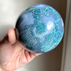 Pu&ograve; includere: Una pietra sferica lucidata con un motivo a spirale di colori blu-grigio e verde-acqua. La gemma &egrave; tenuta in una mano, mostrando la sua forma liscia e arrotondata e le sue variazioni naturali. La superficie riflette la luce, evidenziando i suoi motivi unici.