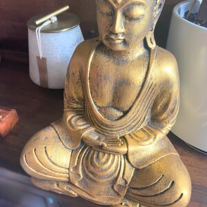 May include: A gold-colored Buddha statue sitting in a meditative pose with hands clasped in front. The statue has a detailed face and intricate folds in the clothing.