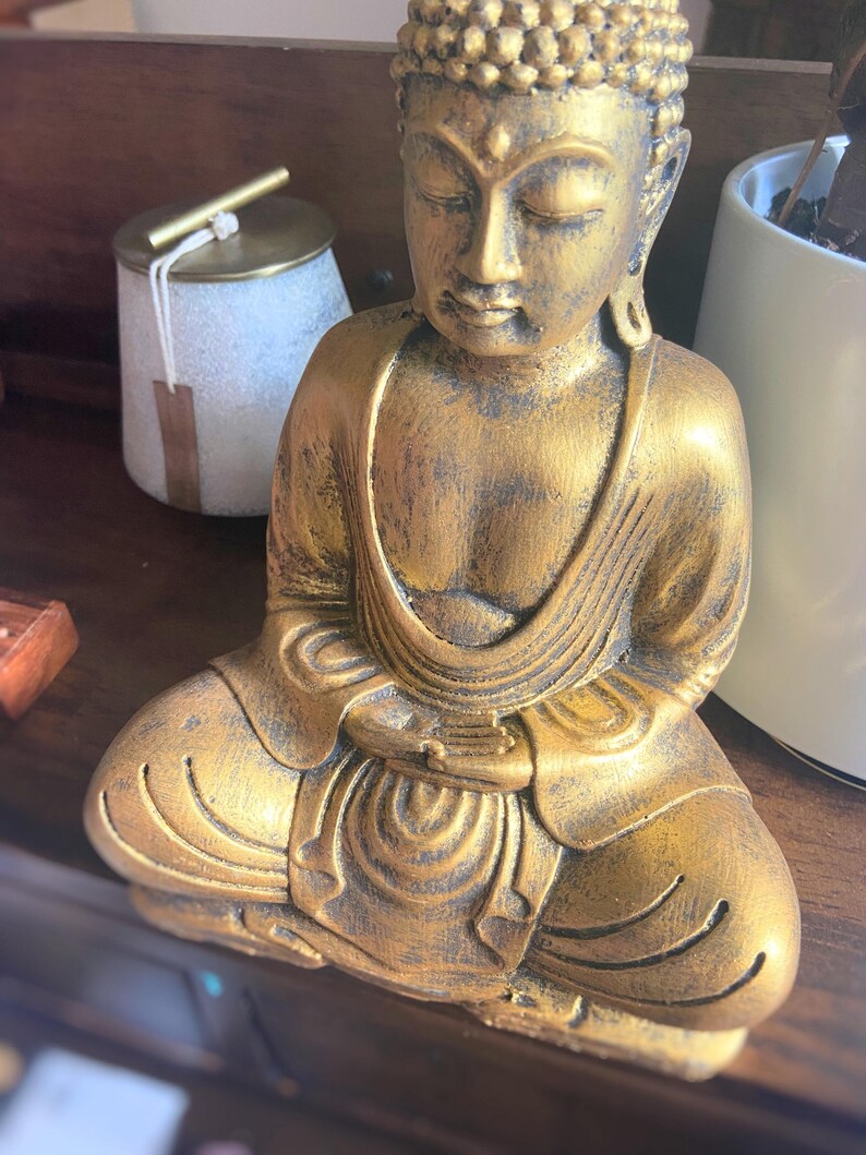May include: A gold-colored Buddha statue sitting in a meditative pose with hands clasped in front. The statue has a detailed face and intricate folds in the clothing.