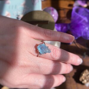 May include: A silver ring featuring a raw, light blue aquamarine gemstone set in a prong setting. The stone has an irregular, jagged shape with a translucent, crystalline texture. The metal band is thin and polished, encircling the finger.