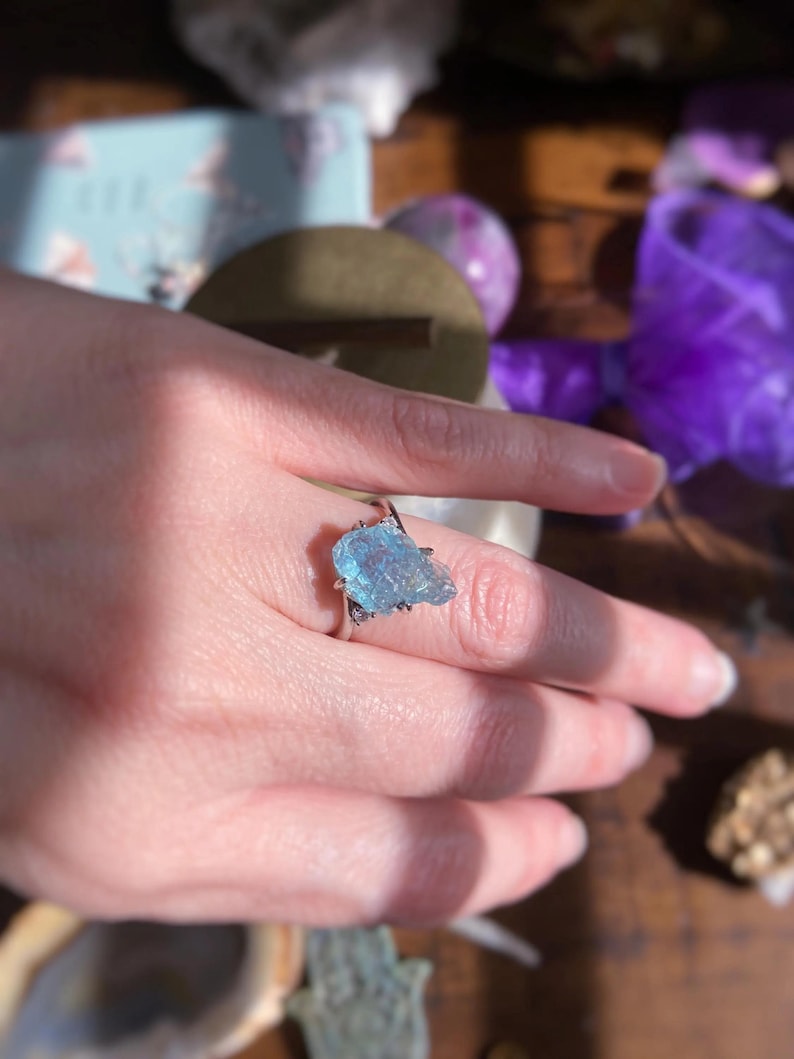 May include: A silver ring featuring a raw, light blue aquamarine gemstone set in a prong setting. The stone has an irregular, jagged shape with a translucent, crystalline texture. The metal band is thin and polished, encircling the finger.