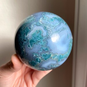 Pu&ograve; includere: Una gemma sferica lucidata con un motivo a vortice di tonalit&agrave; blu e verde acqua. La pietra a forma di sfera ha una superficie liscia e riflettente. La gemma &egrave; tenuta in mano. Un oggetto decorativo.