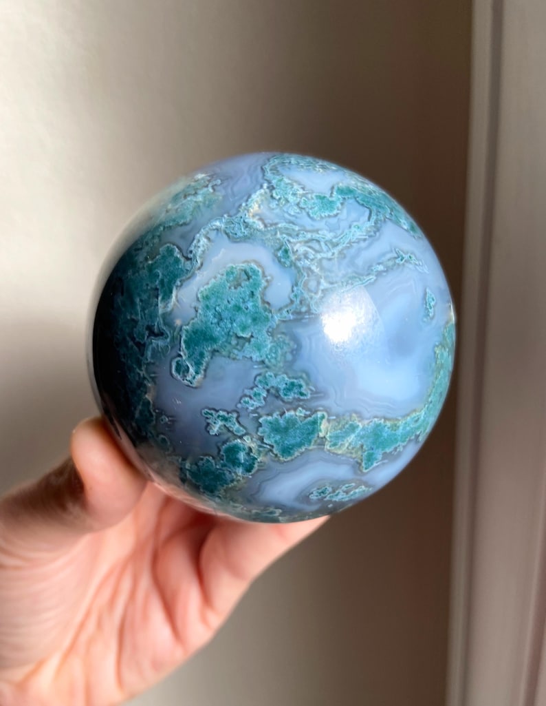 Pu&ograve; includere: Una gemma sferica lucidata con un motivo a vortice di tonalit&agrave; blu e verde acqua. La pietra a forma di sfera ha una superficie liscia e riflettente. La gemma &egrave; tenuta in mano. Un oggetto decorativo.