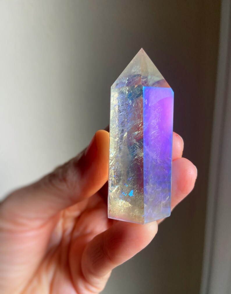 Puede incluir: Una punta de cristal hexagonal con acabado pulido, que muestra una apariencia transl&uacute;cida con fracturas internas y un revestimiento iridiscente vibrante que refleja tonos azules, p&uacute;rpuras y dorados en su superficie facetada.
