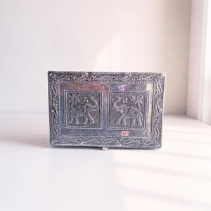 May include: A rectangular silver-toned metal box featuring an embossed design of two elephants standing under palm trees. The lid is framed by a decorative rope-like border with floral motifs, creating a detailed metallic surface finish.
