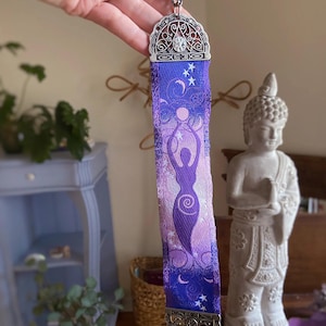 May include: A purple and blue bookmark with a silver filigree top and bottom. The bookmark features a silhouette of a goddess figure, moons, and stars. The bookmark is held up by a hand.