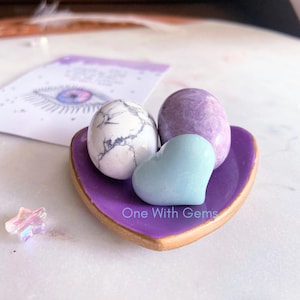 May include: A heart-shaped purple dish with gold trim holds three gemstones: a white and gray speckled egg-shaped stone, a lavender egg-shaped stone, and a light blue heart-shaped stone. A card with an eye illustration is in the background.