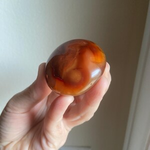 May include: A smooth, polished, rounded carnelian stone featuring a translucent appearance with deep orange, reddish-brown, and amber color gradients. The stone has a glossy, reflective surface with natural internal banding and variations.