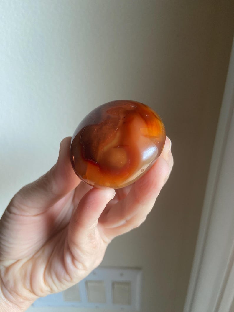 May include: A smooth, polished, rounded carnelian stone featuring a translucent appearance with deep orange, reddish-brown, and amber color gradients. The stone has a glossy, reflective surface with natural internal banding and variations.