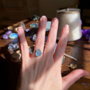 May include: A silver-toned ring featuring a raw, uncut, light blue gemstone set in a prong setting. The stone has a jagged, irregular shape with a translucent appearance. The band of the ring is thin and metallic, encircling the finger.