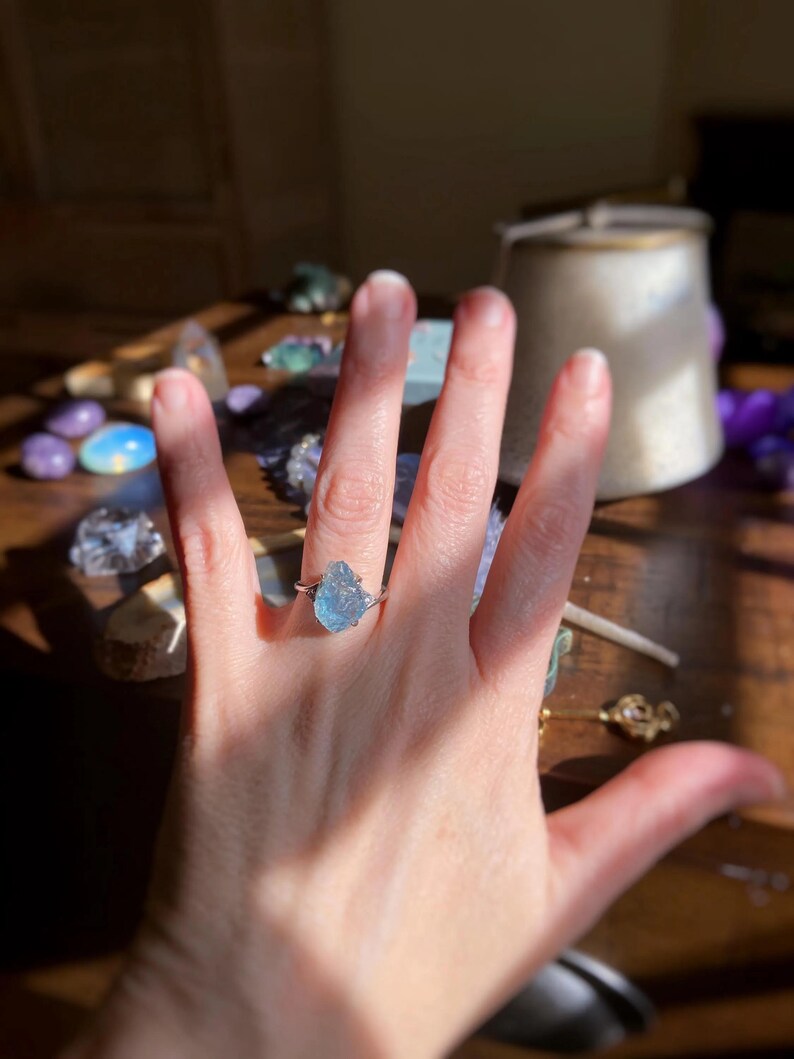 May include: A silver-toned ring featuring a raw, uncut, light blue gemstone set in a prong setting. The stone has a jagged, irregular shape with a translucent appearance. The band of the ring is thin and metallic, encircling the finger.